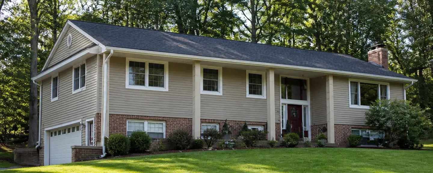 Bi-level home with tan vinyl siding over a brick lower level, a charcoal shingle roof, and tall trees behind it.