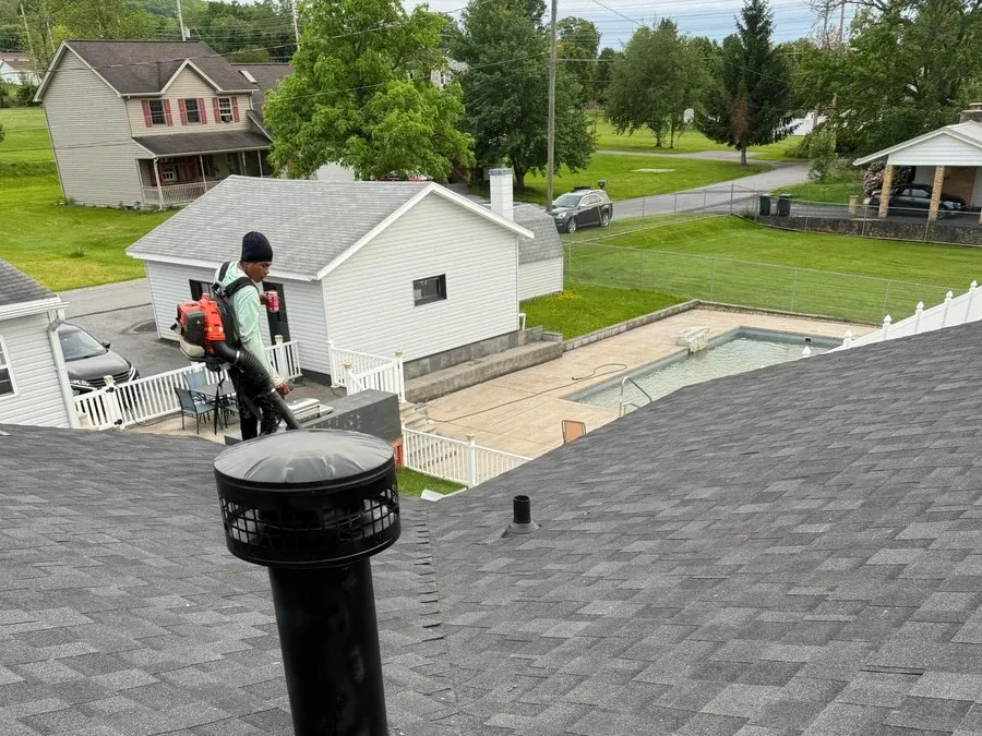 Roofer walking a shingle roof and clearing debris near a plumbing vent boot during an inspection.