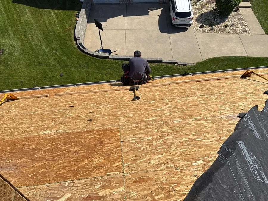 Roofer seated on bare OSB roof decking beside rolled synthetic underlayment and roofing rakes on a new construction project.