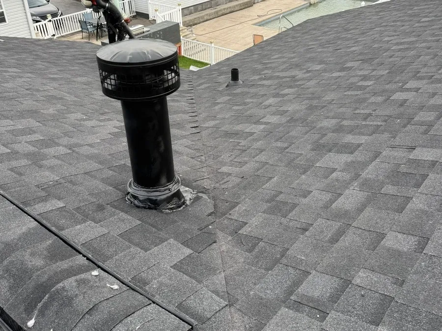 Roofing crew member with a tool working beside a vent pipe boot on a shingle roof, curled underlayment edge visible in the foreground.