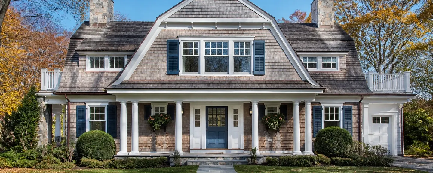 Gambrel-roof American shingle style home with blue shutters and autumn foliage.
